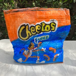 May include: A hand-painted, orange and blue Cheetos Cuffs bag. The bag features the Cheetos logo in yellow and black, with a cartoon image of Chester Cheetah. The bag is crumpled and appears to be made of paper.
