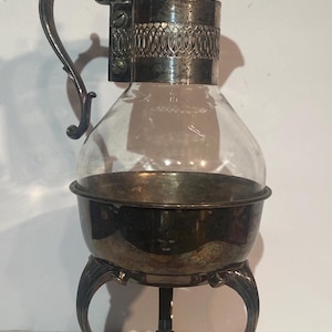 Puede incluir: Un antiguo servidor de café plateado con una jarra de vidrio transparente. El servidor presenta un asa decorativa, una tapa y un soporte con patas ornamentadas. La jarra se sujeta con una banda plateada con un patrón ovalado.