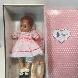 Puede incluir: Un vestido rosa y blanco con un lazo rosa y un borde de encaje blanco. La muñeca tiene el pelo castaño y los ojos azules. La muñeca está en una caja blanca con las palabras "effanbee doll company since 1921" en el lateral.
