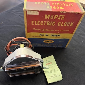 May include: A vintage Mopar electric clock with its original box and paperwork. The box is red and yellow with the text "Mopar Electric Clock" and "Part No. 1300609". The clock is silver and black with a clear face. The paperwork reads "Genuine Borg Electrically Wound Clock."
