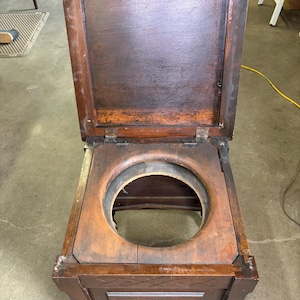 Puede incluir: Una silla de noche antigua de madera con tapa con bisagras y una abertura circular. La silla de noche es de madera teñida oscura y tiene tallas decorativas en el panel frontal. Se apoya sobre pequeñas ruedas.