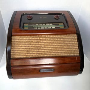 Op de afbeelding: Een Philco tafelradio met een donkere houtnerfafwerking, een gebogen voorpaneel en een luidsprekerrooster van gestructureerde beige stof. Het bovenste gedeelte bevat een zwart bedieningspaneel met twee ronde knoppen en een frequentieschaal.