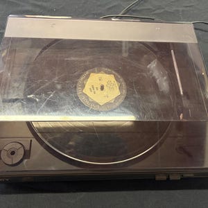 Może przedstawiać: Czarny gramofon Sony z przezroczystą plastikową pokrywą. Płyta winylowa znajduje się na gramofonie. Gramofon posiada przewód zasilający oraz panel sterowania z przyciskami i pokrętłem. Etykieta płyty jest widoczna.