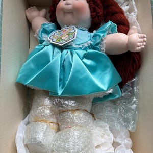 May include: A Cabbage Patch Kids doll with dark red yarn hair, a pale complexion, and a bright turquoise satin dress with white lace trim. The doll features a tag on the chest with the text Cabbage Patch Kids and is packaged in a cardboard box.
