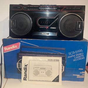 May include: Black Randix SCR-5195 AM/FM stereo cassette recorder with large speaker grilles. The boombox has a built-in radio tuner and cassette player. The original box and manual are included. The box is blue with the Randix logo.