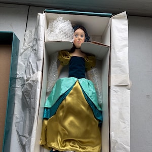 May include: A porcelain doll in a box, wearing a teal and gold dress with a dark blue bodice and yellow sleeves. The doll has dark hair and is packaged with protective materials.