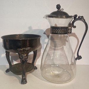 May include: A clear glass carafe with a silver-colored lid and handle, next to a silver-colored bowl on a decorative stand. The carafe has a wide base and a narrow neck. The bowl is round and sits on three ornate legs. The items appear to be antique.