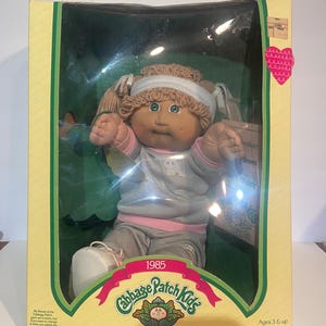 Könnte beinhalten: Eine Cabbage Patch Kids Puppe aus dem Jahr 1985 in der Originalverpackung. Die Puppe hat Wollhaare, grüne Augen und trägt ein graues Langarmshirt mit rosa Besatz, eine graue Hose und weiße Schuhe. Die Schachtel zeigt den Text Cabbage Patch Kids und I'm unique. I'm adoptable!