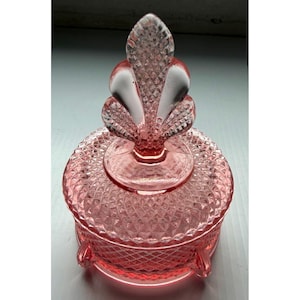 Op de afbeelding: Een ronde roze glazen sieradendoos met een ruitpatroon op het deksel en de basis. Het deksel is bekroond met een decoratieve knop in de vorm van een gestileerd blad of een lelie, en de basis rust op drie kleine, korte pootjes.