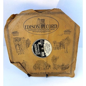 May include: A vintage Edison Record paper sleeve in tan, featuring black printed illustrations of musicians and singers. The text reads Edison Record, Comparison with the Living Artist Reveals No Difference, and Thomas A. Edison Inc., Orange, N.J., U.S.A.