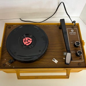 May include: A vintage Symphonic Solid State record player in a yellow case with a brown wood-grain top. The turntable has a black platter with a red 45 RPM adapter. Controls include volume and tone knobs.