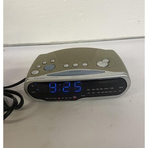Könnte beinhalten: Ein silberner und grauer digitaler Radiowecker mit einer blauen LED-Anzeige, die 9:25 anzeigt. Das Gerät verfügt über ein oben angebrachtes Lautsprechergitter, verschiedene Bedientasten und eine Schlummertaste. Die Frontplatte zeigt SmartSet.