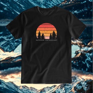 Può includere: T-shirt nera con un design a tramonto nei toni dell'arancione, rosso e giallo, e sagome di pini. Il testo "OUTDOOR ENTHUSIAST" è stampato sotto. Lo sfondo mostra montagne e un lago.