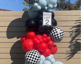 May include: A balloon arrangement featuring black, red, and light blue balloons, with checkered pattern balloons. A sign reading "SPEED LIMIT 2" is centered in the arrangement. The balloons are arranged in a vertical column.