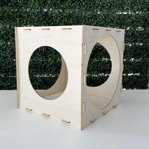 May include: A light-colored wooden cube with two large circular cutouts. The cube has the text "7" and "10" on its sides, indicating measurements in inches. The cube is constructed with interlocking joints and sits on a white surface.