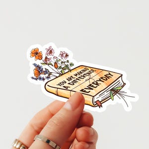 May include: A sticker featuring a watercolour illustration of a book with the text "YOU ARE MAKING A DIFFERENCE EVERYDAY." The book is adorned with colourful flowers. The sticker is held by a hand.