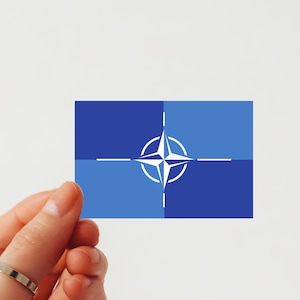 Peut inclure: Un autocollant rectangulaire avec le drapeau de l'OTAN. Le drapeau est divisé en quatre sections bleues, avec un emblème de rose des vents blanc au centre. L'autocollant est tenu sur un fond blanc.