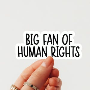 May include: A white sticker with the text "BIG FAN OF HUMAN RIGHTS" in black, sans-serif font. The sticker is held by a hand with rings on the fingers. The background is plain white.
