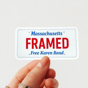 Puede incluir: Una pegatina blanca con forma de matrícula de Massachusetts. La pegatina tiene las palabras "Massachusetts" en azul, "FRAMED" en rojo y "Free Karen Read" en azul. La pegatina está sujeta por una mano.