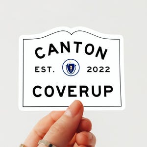 May include: A white sticker with a black border and the words "CANTON COVERUP" in a bold, sans-serif font. The sticker also includes "EST. 2022" and a circular emblem. The sticker is held by a hand.