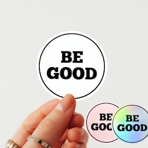 May include: White circular sticker with the words "BE GOOD" in bold black font, encircled by a black outline. Two additional stickers are visible, one pink and one holographic, each with the same text. The stickers are held by a hand.