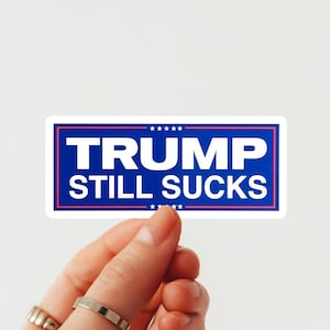 May include: A rectangular sticker with the text "TRUMP STILL SUCKS" in white letters on a blue background, framed by a pink border. The sticker is held by a hand against a white background.