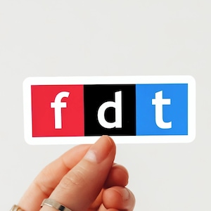 Puede incluir: Una pegatina rectangular con las letras blancas "fdt" sobre bloques rojos, negros y azules. La pegatina está sujeta por una mano sobre un fondo blanco.