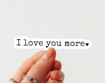May include: A white rectangular sticker with a die-cut border featuring the black typewriter-style text I love you more followed by a small black heart symbol. The sticker has a clean white background and clear, bold lettering.