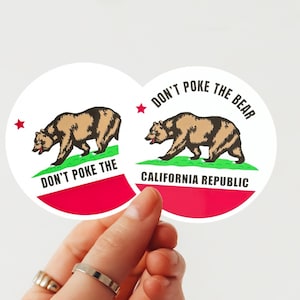 May include: Two round stickers featuring the California state flag design. Each sticker displays a brown bear walking on green grass, with the text "DON'T POKE THE BEAR" and "CALIFORNIA REPUBLIC". The stickers have a red and white background.