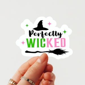Könnte beinhalten: Ein weißer Aufkleber mit dem Schriftzug „Perfectly WICKED“ in Schwarz, Grün und Pink, mit einer Hexenhut- und Besen-Grafik. Grüne und rosa Sternchen akzentuieren das Design. Der Aufkleber wird von einer Hand gehalten.