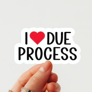 Pode incluir: Um autocolante branco com o texto "I ❤️ DUE PROCESS" em preto, com um coração vermelho a substituir a palavra "love". O autocolante é segurado por uma mão contra um fundo branco.