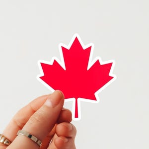 May include: A red maple leaf sticker with a white border is held between fingers against a white background. The maple leaf is a symbol of Canada. The sticker is a simple, graphic design.