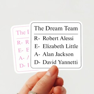 Puede incluir: Dos pegatinas blancas con texto negro. Una dice "The Dream Team" con una lista de nombres. La otra, en rosa, dice "The I READ". Las pegatinas están sujetas por una mano.