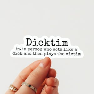 May include: A white sticker with the word "Dicktim" in bold black letters. Below, a definition reads: "(n.) a person who acts like a dick and then plays the victim." The sticker is held by a hand with rings.
