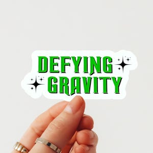 May include: A white sticker with the words "DEFYING GRAVITY" in green, outlined in black. The sticker has a white border and is decorated with black starbursts. The sticker is held by a hand.