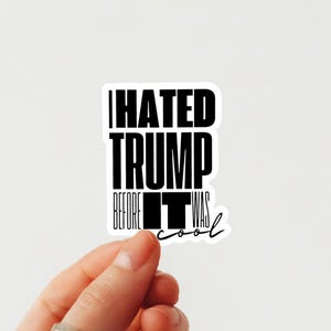 Pode incluir: Um adesivo branco com texto preto que diz "I HATED TRUMP BEFORE IT WAS cool." O adesivo é segurado por uma mão contra um fundo branco. O texto está em negrito, fonte sem serifa.