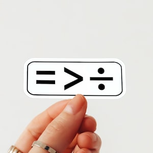 May include: A white sticker with a black border featuring mathematical symbols: an equals sign, a greater-than sign, and a division symbol. The sticker is held by a hand with rings.