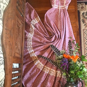 Può includere: Un abito maxi vintage rosa a fiori con bordino in pizzo bianco e schiena con lacci. L'abito è drappeggiato su una testiera in legno e in primo piano c'è un'arrangiamento floreale in una teiera d'argento.