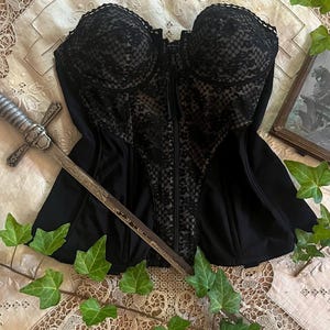 May include: Black corset with lace detailing and a front zipper. A decorative sword rests across the corset. The corset is displayed on a vintage, cream-colored lace tablecloth, with green ivy accents.