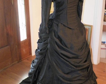Puede incluir: Un vestido negro de estilo victoriano con un corpiño ajustado y una falda completa. El vestido tiene un escote con volantes y mangas largas. La falda está fruncida y tiene un volante escalonado en el dobladillo.
