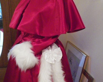 May include: A red satin dress with a white fur trim and a white satin bow. The dress has a cape-like top and a long skirt.
