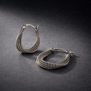 May include: A pair of silver-toned hoop earrings. Each earring features a textured, woven design, with a curved shape. The earrings are displayed on a dark gray surface, with the light reflecting off the metal.