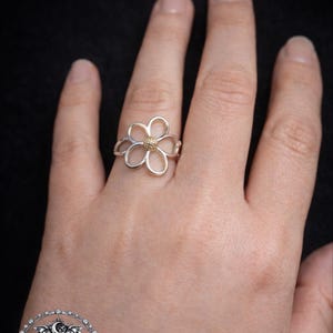 Peut inclure: Une bague fleur argentée avec un centre doré est portée sur une main. La bague présente un motif de fleur ajourée. Le fond est noir uni, ce qui met en valeur les détails de la bague. C'est un bijou.