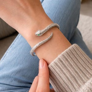 Puede incluir: Una pulsera de serpiente plateada con un patrón de escamas texturizadas. La cabeza de la serpiente tiene un ojo de piedra verde. La pulsera se envuelve alrededor de la muñeca, creando un accesorio elegante. La pulsera se muestra en una muñeca.