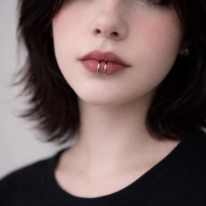 May include: Close-up of a person's lips with two silver lip rings. The person has dark hair and is wearing a black shirt. The lips are painted with a mauve lipstick.