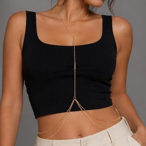 Pode incluir: Colar de corrente dourada para o corpo. O colar apresenta uma corrente delicada que cai do pescoço, sobre o peito e ao redor da cintura. A joia corporal é usada sobre uma regata preta e calças bege.