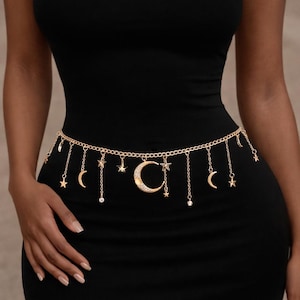Puede incluir: Cinturón de cadena dorado con colgantes. El cinturón presenta pequeñas estrellas, medias lunas y un gran colgante de media luna. Se lleva sobre un vestido negro.