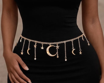 May include: Gold-colored chain belt with dangling charms. The belt features a series of small stars, crescent moons, and a large crescent moon pendant. The belt is worn over a black dress.