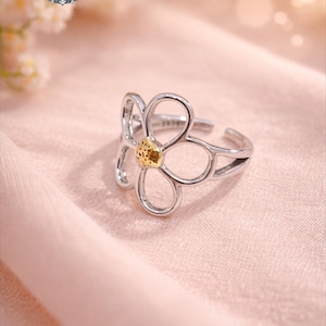 Peut inclure: Une bague fleur argentée avec un centre jaune, sur un fond rose tendre. La bague présente un design ajouré avec cinq pétales. La bague est réglable et porte le nom de la marque "NovaCraft Cie" dans le coin supérieur gauche.