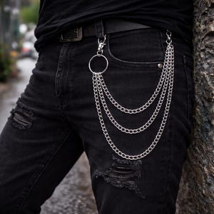 Puede incluir: Cadena de billetera plateada con tres hebras, sujeta a una trabilla con un anillo grande y un cierre. La cadena cuelga sobre el bolsillo delantero de unos vaqueros negros desgastados. Los vaqueros se llevan con un cinturón negro y una camisa negra.